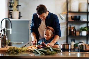 Smiling father showing his cute little daughter how to cut vegetables while preparing a healthy meal at home