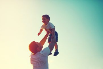 father lifting his son overhead with green and yellow gradient sky behind them